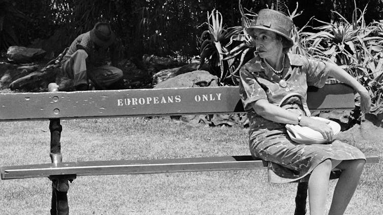 A black and white photo of a park. A caucasian woman in a hat clutches her handbag as she sits on a bench that has a sign reading Europeans Only. In the background an African man crouches as he tends the garden.