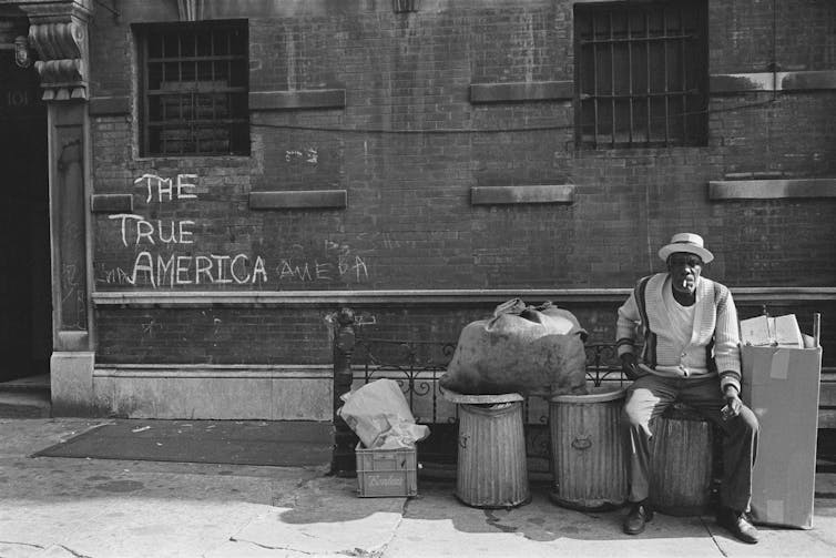 A black and white photo of an elderly African American man in a hat. He sits on a garbage can, a cigarette drooping from his lip, and looks frankly at the camera. Graffitti on the brick wall behind him reads, The True America.