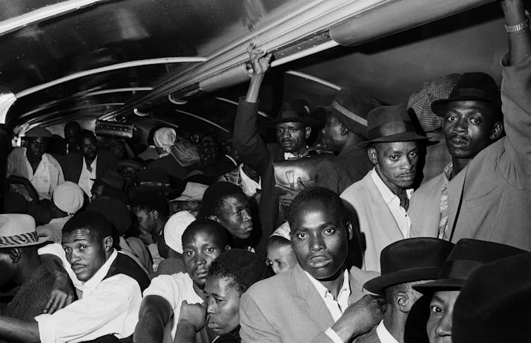 A black and white photo of a group of men is packed into a train, holding on where they can, their expressions blank or grim.
