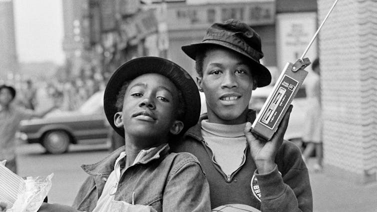 A black and white photo of two young African American boys in hats. One holds a radio in the air as they smile and pose for the camera, full of mischief.