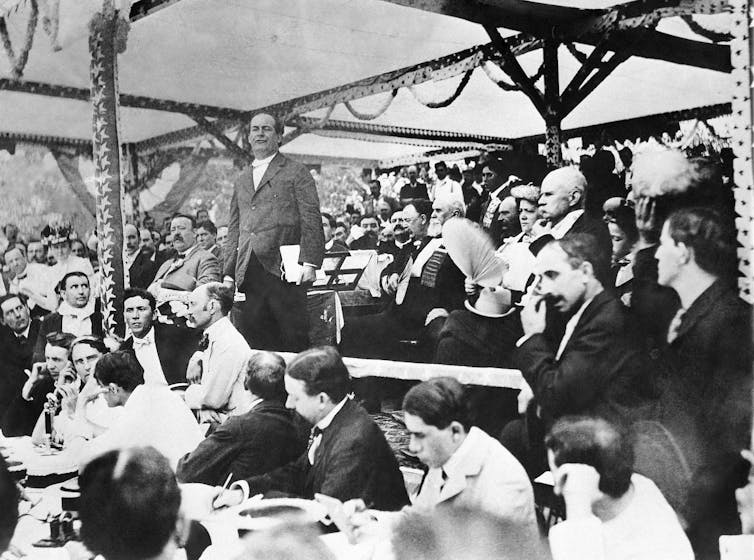 A man in a gray suit stands and speaks to a crowd of seated people.