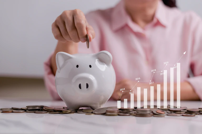 A person, seen from the neck down, puts a coin in a piggy bank. A visual of a bar graph increasing in value over time is seen beside it.