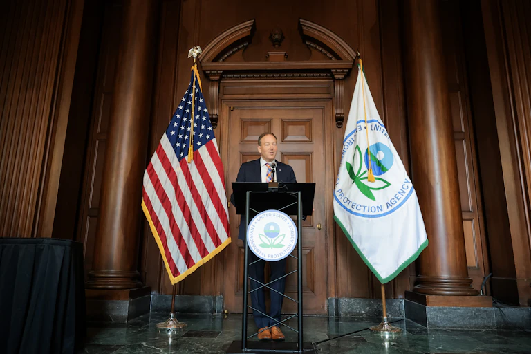 A man in a suit stands at a lectern; behind him are a U.S. flag and the flag of the U.S. Environmental Protection Agency.
