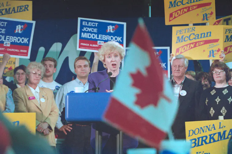 A women with short, wavy blonde hair in a blue blazer speaks from behind a podium to a group of people holding signs supporting the Progressive Conservation party