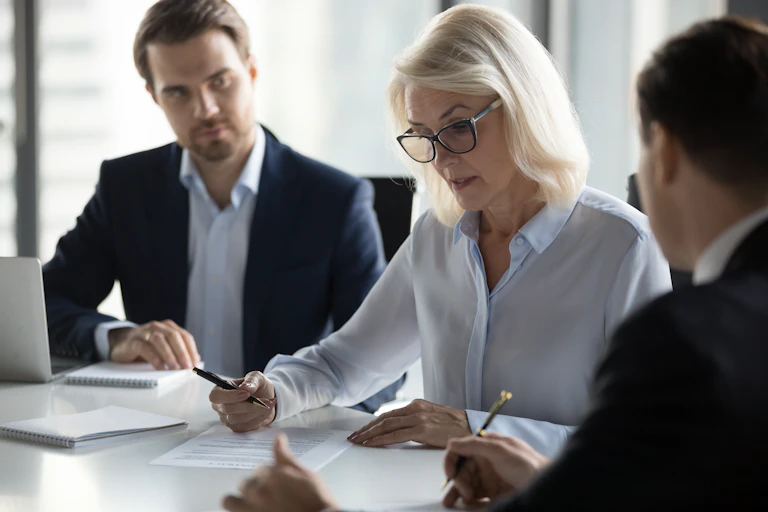 Two men in suits look at a women, who is wearing a button down, as she speaks while writing on a document