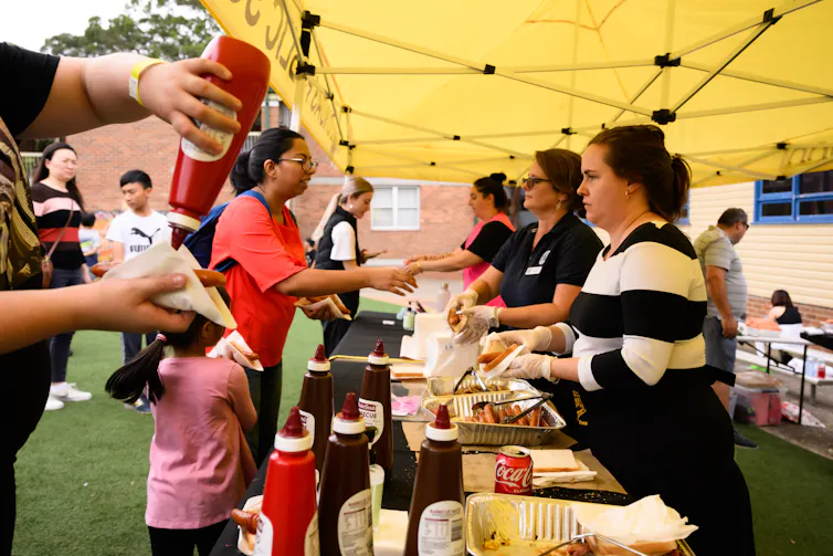 Woman in black and striped jumper serving a sausage sandwich to a woman in a red top and child dressed in pink. Another person squeezes a red tomato sauce bottle above an open sandwich