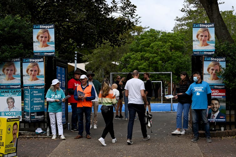 Several large political corflutes framing an entrance to a polling station. Three people wearing colorful, party t-shirts handing out how to vote cards to two people walking past