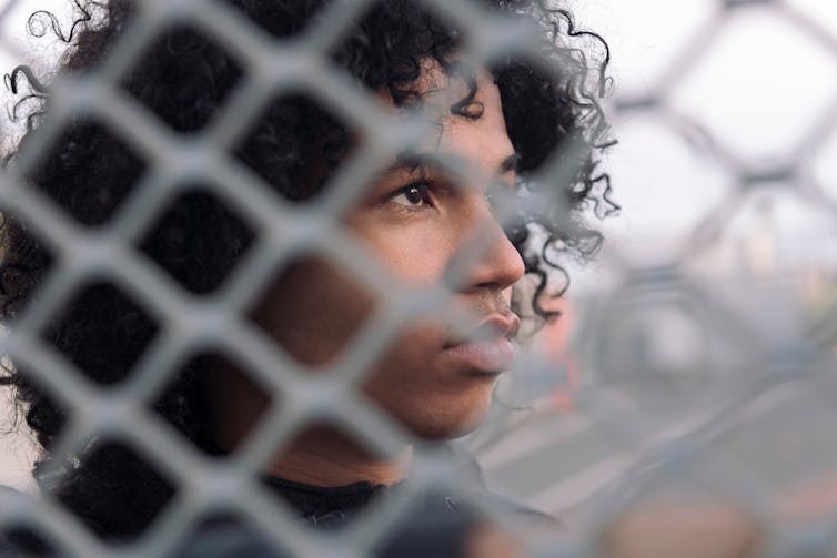 Teen boy looks pensive, seen through metal fence grid.