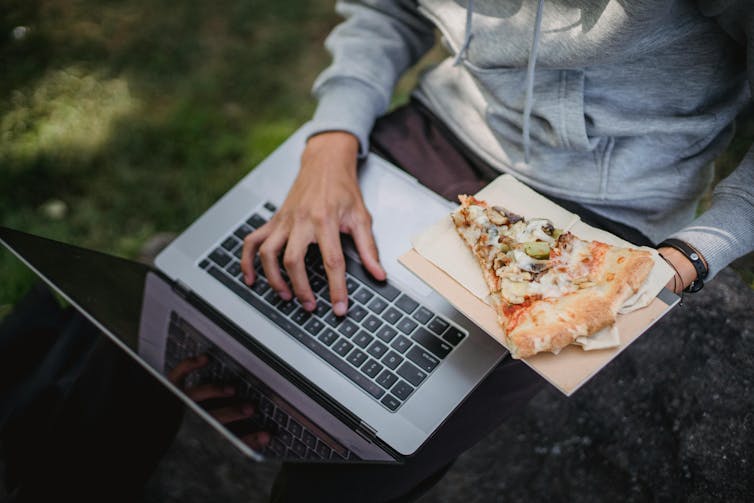 A young person types on a laptop while holding a slice of pizza.