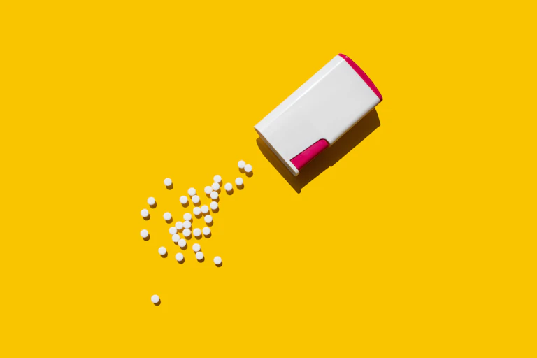 A box of artificial sweetener tablets on a yellow background. A handful of these sweetener tablets are clustered near the box.
