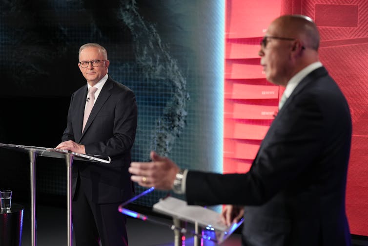 Two men stand at lecterns in front of a red and blue backdrop
