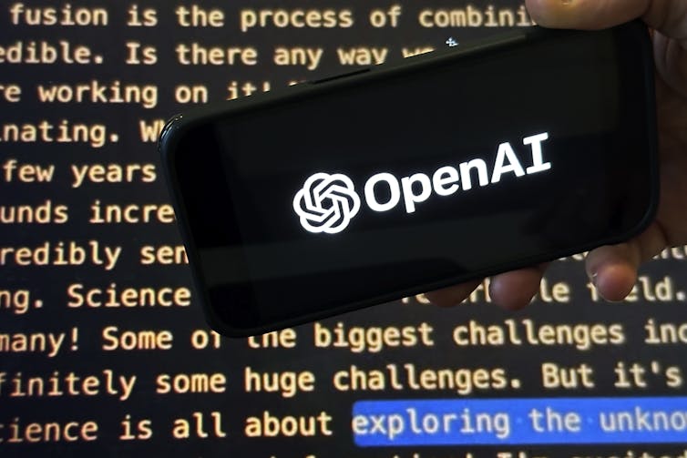 The OpenAI logo appears on a mobile phone in front of a screen showing part of the company website in a photo taken in November 2023