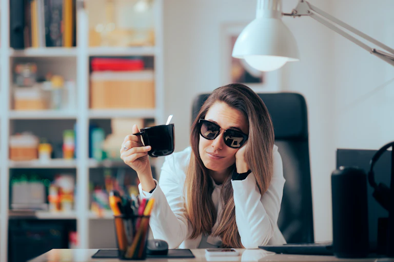 Une femme au travail avec des lunettes de soleil et un mug à la main, qui semble très peu motivée.