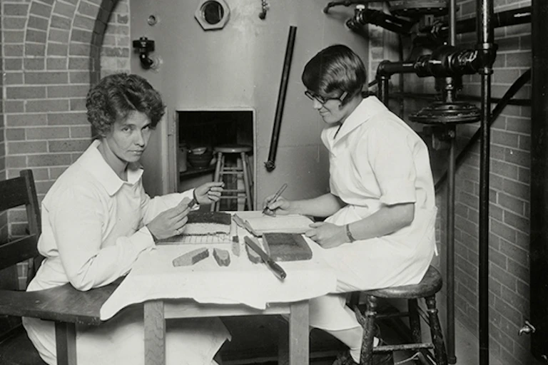 Two women sit at a table in a laboratory wearing white clothing