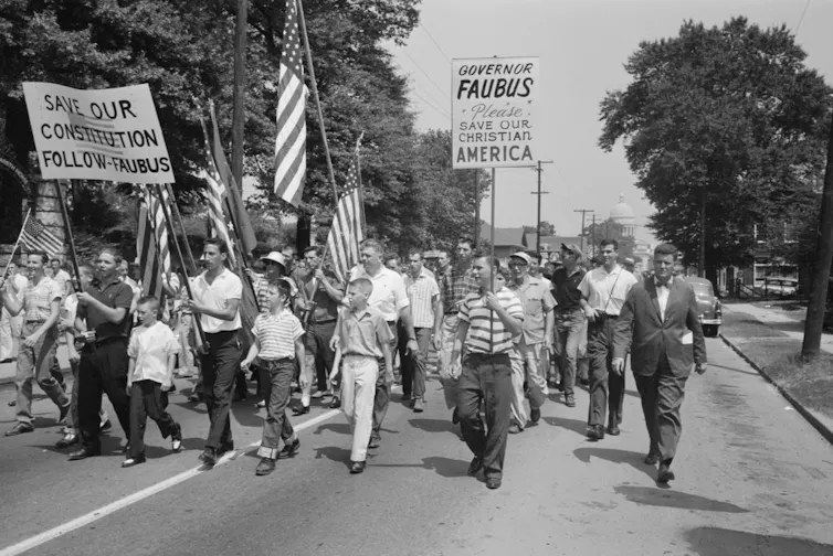 A black-and-white photo of men and boys marching down a street, holding placards and American flags.