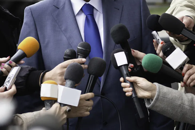 About a dozen microphones are held out toward a man in a suit and tie, whose face is outside the frame of the photo.