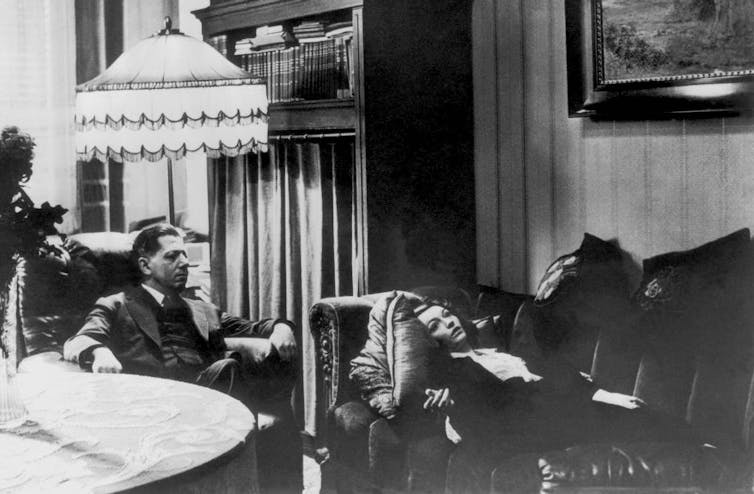 Black-and-white photograph of woman reclining on a couch while a man in a suit sits behind her in a chair.