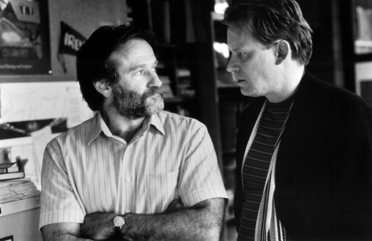 Bearded man with his arms folded speaks to another man.