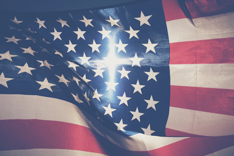 An American flag fills the screen, with a cross-shaped beam of light in the center of it.