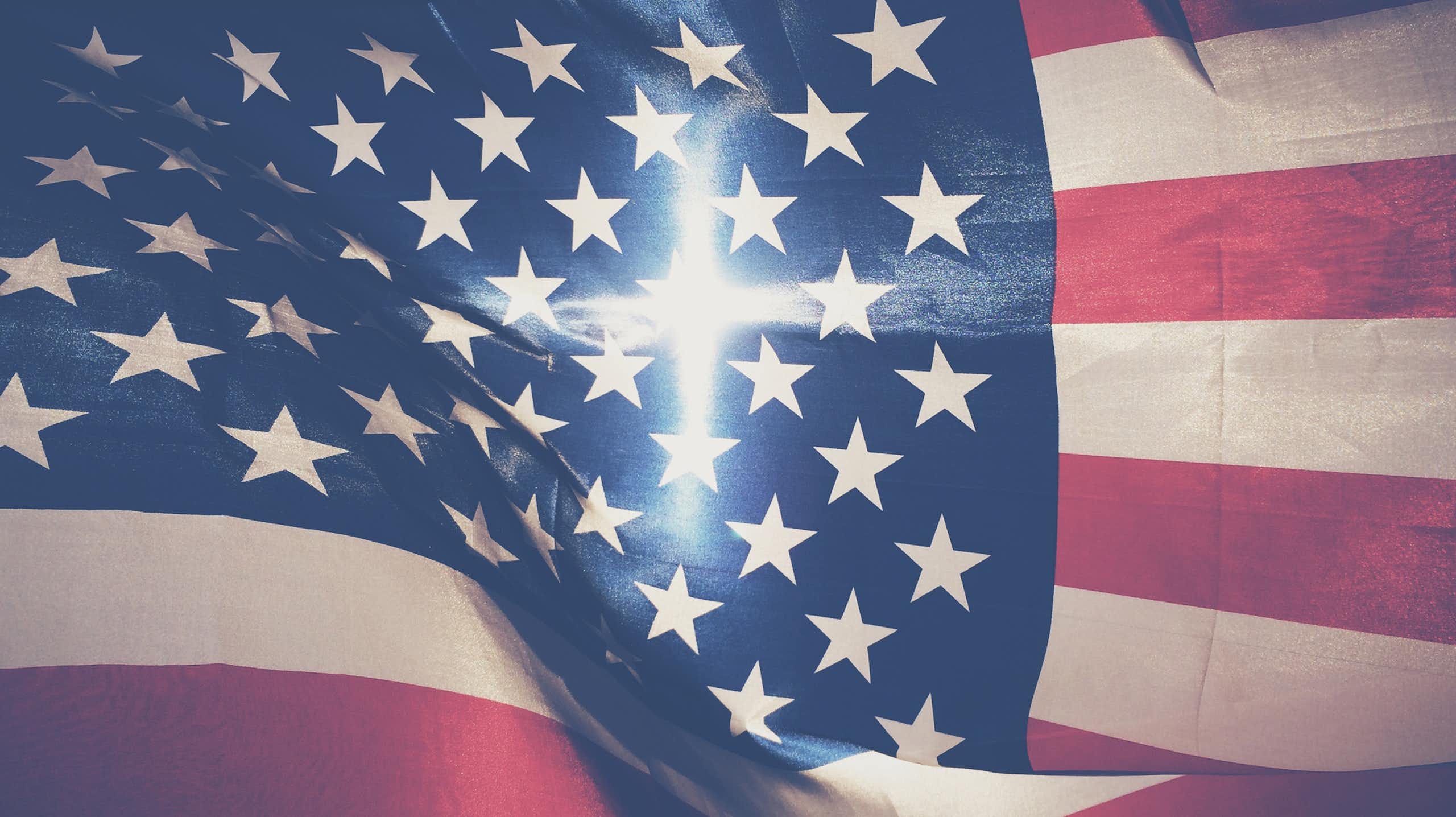 An American flag fills the screen, with a cross-shaped beam of light in the center of it.