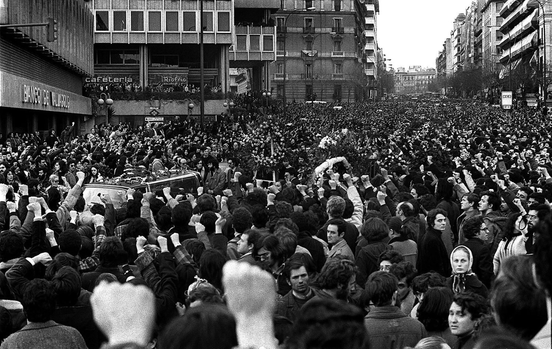 Una foto de la calle llena de personas con sus puños levantados en el paso de los autos funerarios.