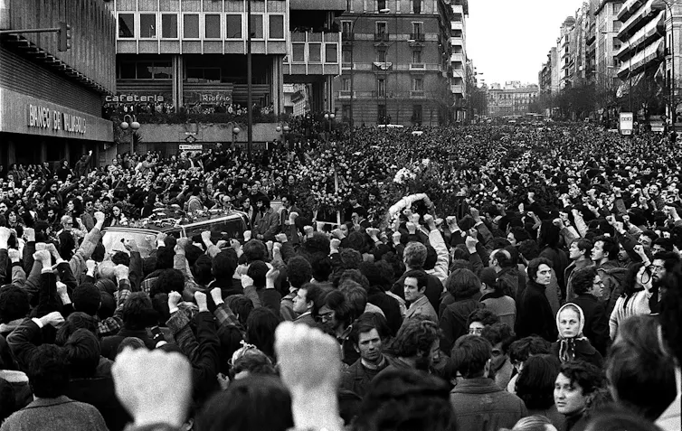 Fotografía de una calle llena de gente con los puños en alto al paso de unos coches fúnebres.