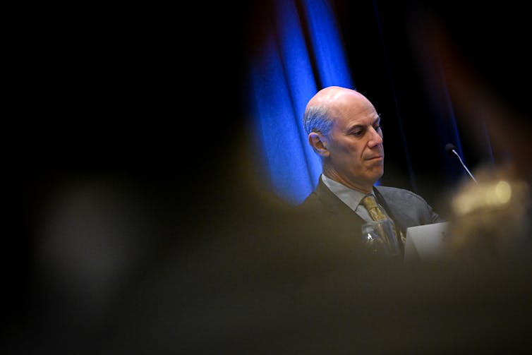 A middle aged, bald man wearing a grey suit is seen looking down in a partially obscured photo.
