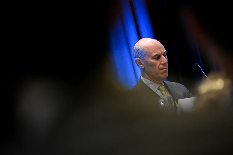 A middle aged, bald man wearing a grey suit is seen looking down in a partially obscured photo.