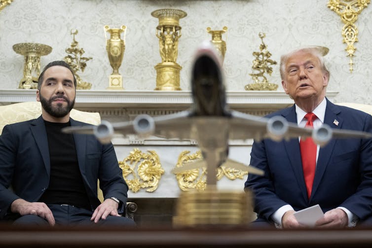 A man with a black beard and a dark blazer and black shirt sits next to an older man with white hair, a blue suit and a red tie. Between them is a model airplane on a table.