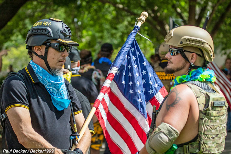 Two men wearing sunglasses and helmets stare at each other while holding the American flag between them.