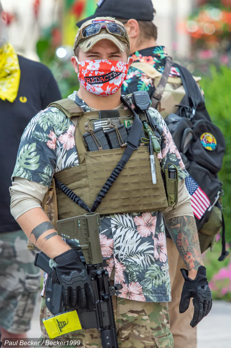 A man with a tattoo on one arm is holding a gun in the other while covering his face with a mask.