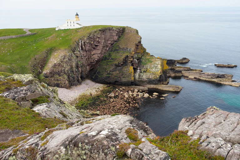 A dramatic eroded cliff in the foreground with a small white lighthouse perched in the distance.
