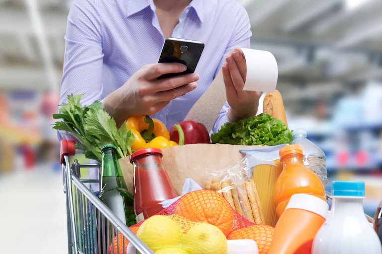 Woman with a loaded shopping trolley checks the grocery receipt and shopping list using her smartphone