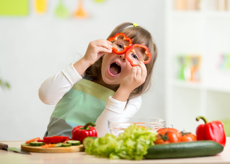 Young girl playing with food, looking through two round slices of capsicum, while preparing fresh vegetables at home in the kitchen.