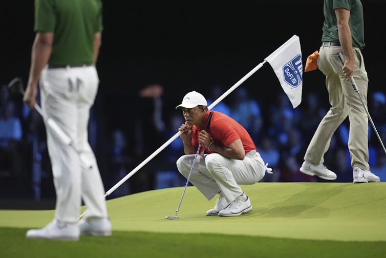 Un hombre con la camisa del polo rojo y las sentadillas de pantalones blancos en el verde de golf mientras sostiene un club de golf en una mano y bandera en otra