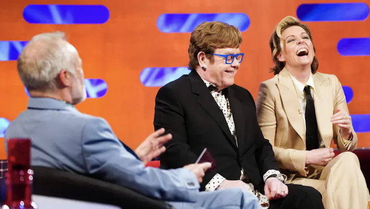 Singers Elton John and Brandi Carlile laughing on a TV chatshow studio set.