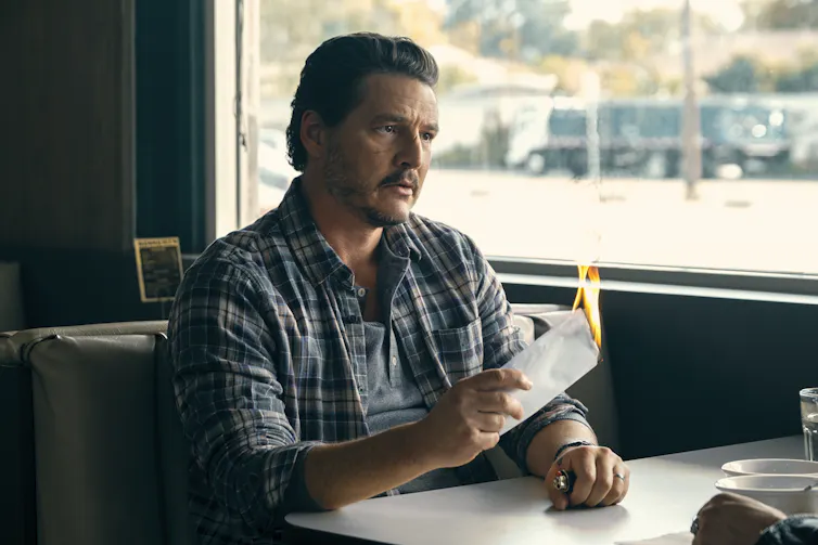 Pedro Pascal sitting in a booth with a flaming piece of paper in his hand