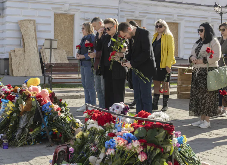 Ukrainians stand in a group with their heads bowed and floral tributes in the foreground.