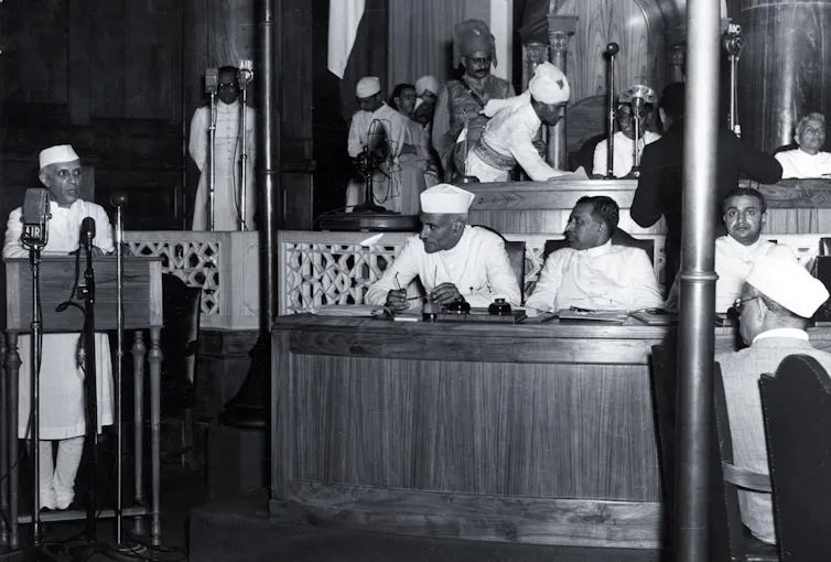 Jawaharlal Nehru, the first prime minister of India, speaks at a podium in the Indian parliament in Delhi, wearing a traditional Indian white coat and hat. Other parliamentarians listen to his speech.