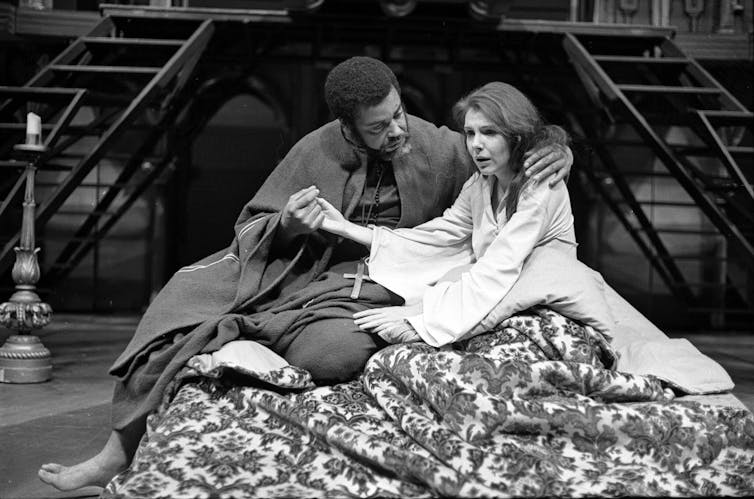 A man and woman hold hands, looking upset, as they sit on a cushion on stage.