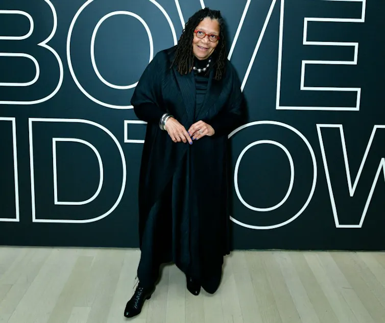 Black woman in a black outfit poses before a black backdrop with white text.