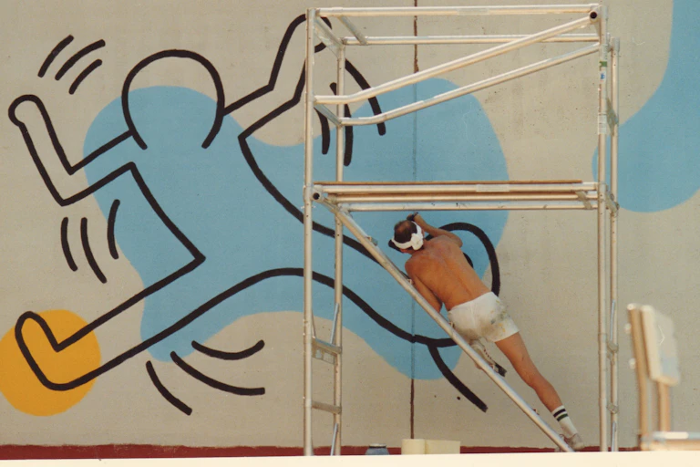 Man paints a large blue and yellow outline of a cartoon person on a wall.