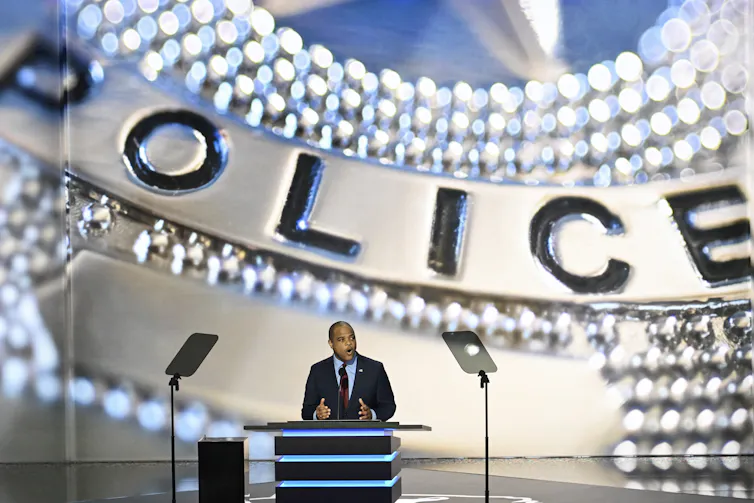 A man at a lectern in a large room with the word 'POLICE' on the wall behind him.