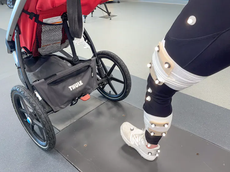 Uma pessoa segurando um carrinho de bebê e correndo para a frente, com a foto focalizando a perna dianteira quando ela atinge o chão. A perna está coberta de pontos brancos e tem um pano enrolado em volta dela para captura de movimento.