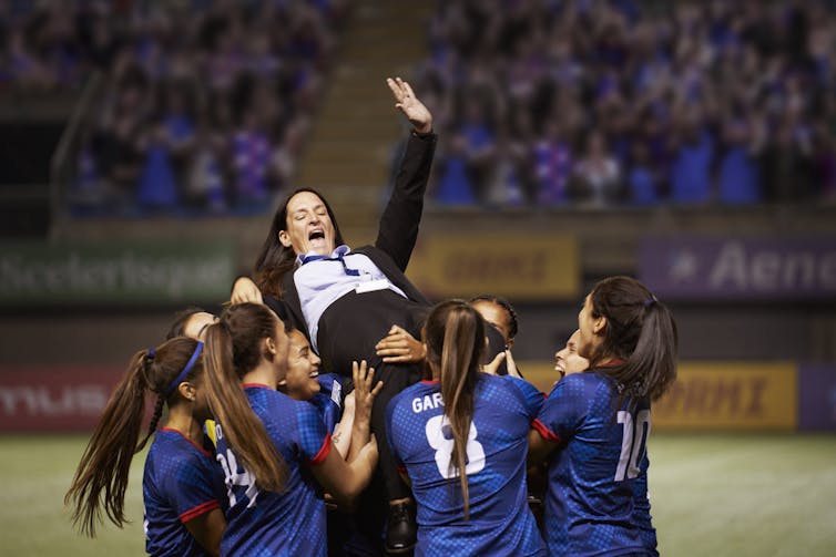 A team of soccer players lift their coach into the air, as she smiles and high fives the air.