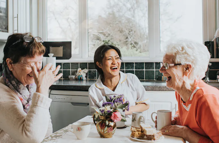 A middle-aged woman sits at a kitchen table between two older women, all of whom are laughing joyously.