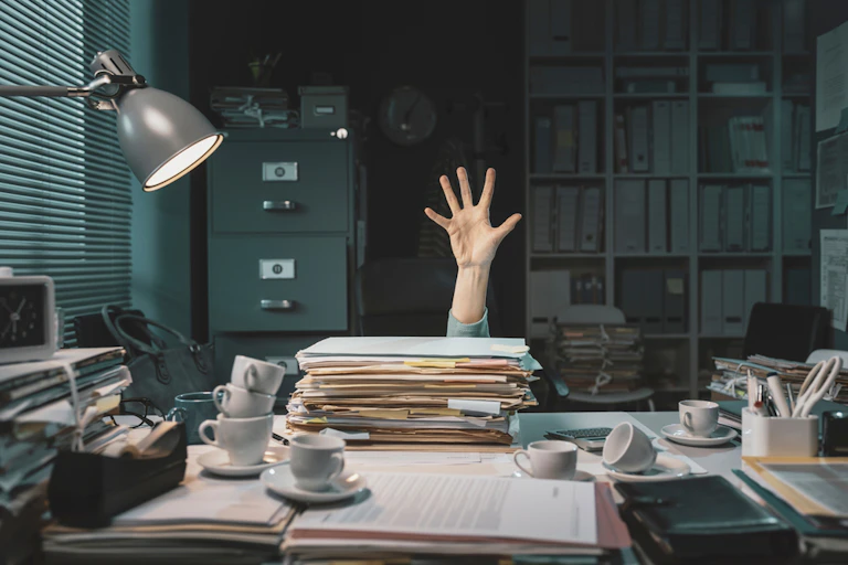 Une personne derrière un bureau au décor vieillot. Elle lève la main comme une personne en train de se noyer signifiant une grande menace.