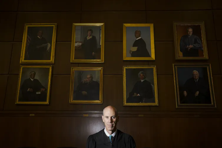 A balding man in judge's robes standing in front of a wall that bears many portraits of judges in gold frames.