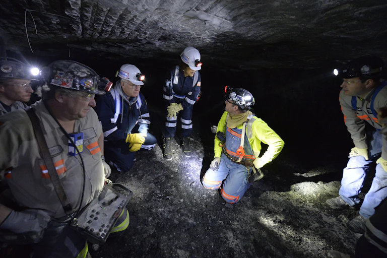 People wearing protective clothing, including hard hats, crouch and kneel in a confined space.