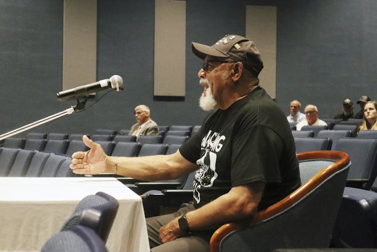 A man sits at a table and speaks into a microphone.